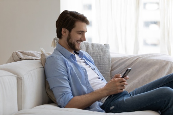 Ein Mann sitzt mit dem Handy in der Hand auf dem Sofa und tippt eine Nachricht.